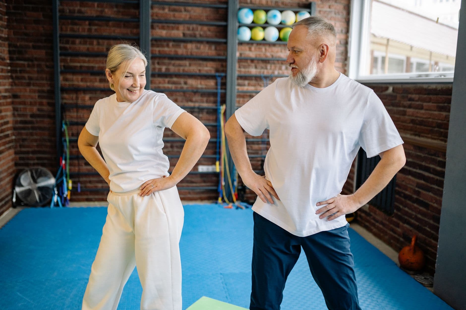 Une femme d'environ 60 ans souriante, en tenue de sport, tenant une chaise pour s'équilibrer lors d'un exercice à domicile.