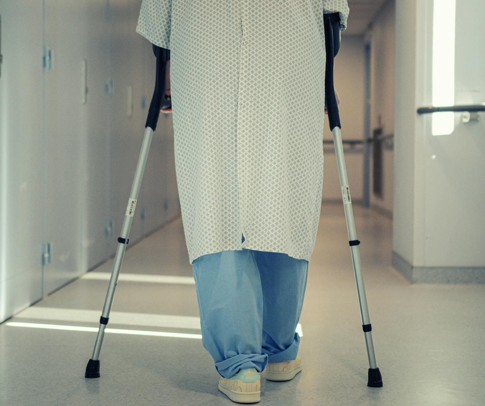 Personne senior marchant avec déambulateur dans un couloir lumineux, accompagnée par un kinésithérapeute