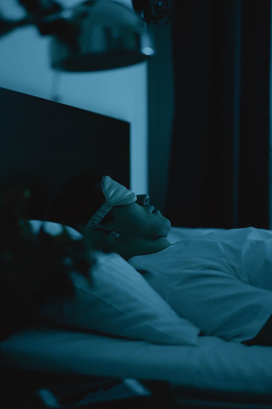 Une personne âgée de plus de 50 ans, allongée paisiblement dans un lit avec une lumière tamisée, les yeux fermés, entourée d'oreillers confortables et d'une couverture douce.