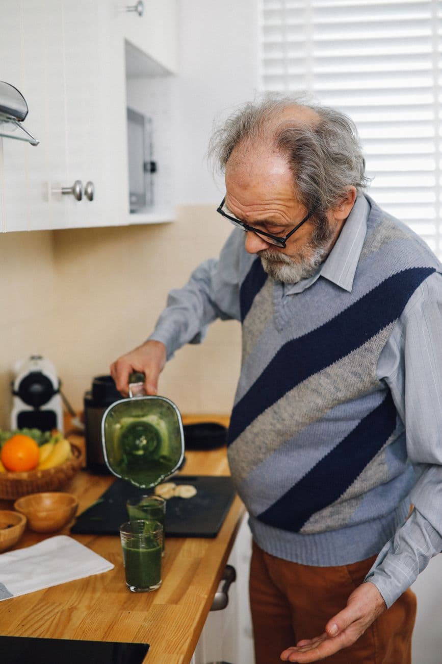 Une personne senior souriante préparant un repas coloré et équilibré dans une cuisine lumineuse, avec des légumes frais, des céréales complètes et des protéines.