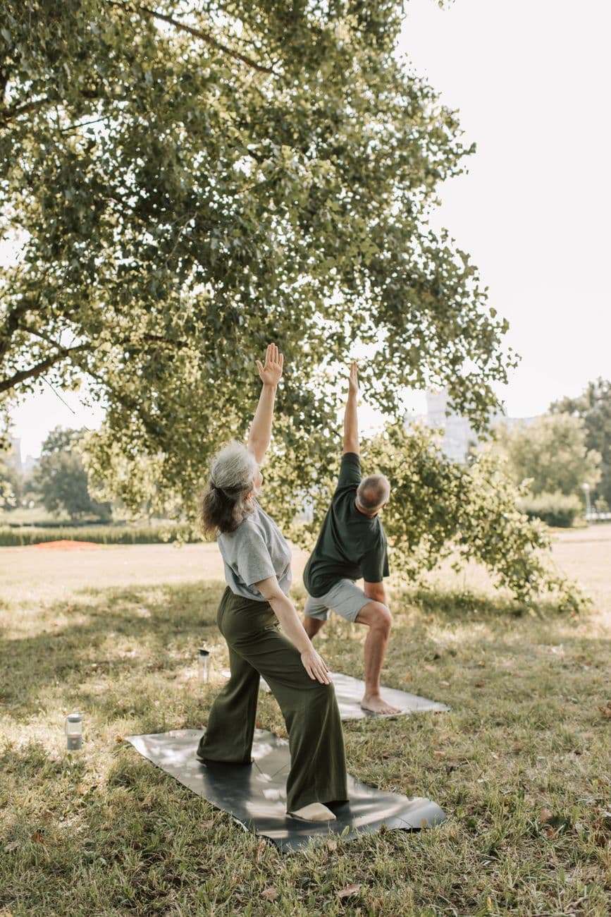 Une femme senior souriante pratique une posture de yoga sur chaise dans un salon lumineux, les bras tendus vers le ciel.