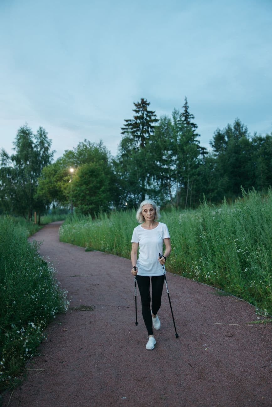 Deux personnes âgées souriantes pratiquant la marche nordique dans un parc, avec des bâtons adaptés, sous un ciel ensoleillé. Elles semblent détendues et heureuses.